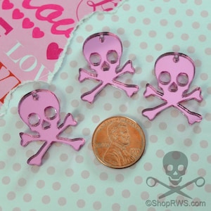 Può includere: Tre ciondoli a forma di teschio e ossa incrociate in specchio rosa. I ciondoli sono circa delle dimensioni di una moneta da un centesimo di euro.