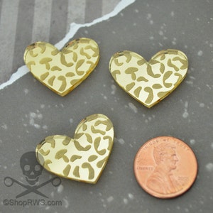 Op de afbeelding: Drie gouden, hartvormige acrylcharms met een luipaardprint. De charms zijn ongeveer zo groot als een Nederlandse cent.