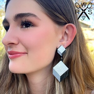 Puede incluir: Un par de pendientes geométricos plateados con un acabado brillante. Los pendientes tienen forma de cubos y están conectados por un hilo negro.