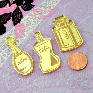 May include: Three gold-colored perfume bottle shaped acrylic charms with the words "adore", "Love Potion", and "Lust" on them. The charms are on a pink and white patterned surface.