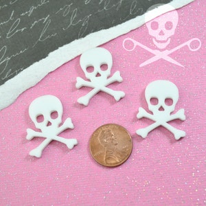 Può includere: Tre ciondoli in plastica bianca a forma di teschio e ossa incrociate su uno sfondo rosa scintillante. I ciondoli sono circa delle dimensioni di una moneta da un centesimo di euro.