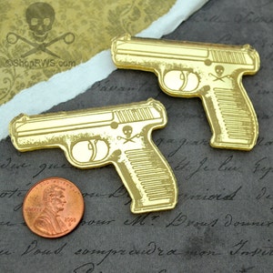 Peut inclure: Deux breloques en plastique en forme de pistolet de couleur dorée avec un motif de crâne et d'os croisés. Les breloques sont sur une surface texturée.
