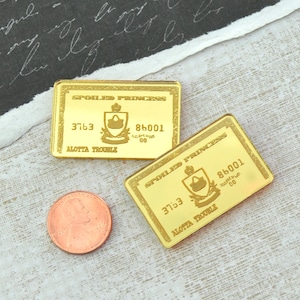 Puede incluir: Dos objetos rectangulares dorados con el texto "SPOILED PRINCESS" y "ALOTTA TROUBLE". Cada objeto tiene un emblema de corona y bolso. Una moneda de cobre está al lado de los objetos. Los objetos están sobre una superficie texturizada.