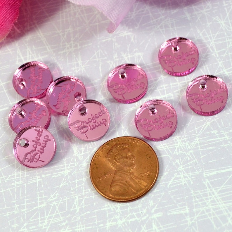Puede incluir: Encantos redondos de acr&iacute;lico rosa con el texto "Project Runway" en letras blancas. Los encantos son aproximadamente del tama&ntilde;o de una moneda de un centavo de euro.