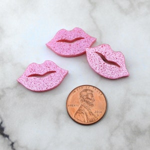 Peut inclure: Trois embellissements en forme de lèvres roses et scintillantes. Une pièce de monnaie américaine est à côté des lèvres pour la comparaison des tailles. Les lèvres sont de couleur rose clair avec une texture scintillante. La pièce est datée de 2014.