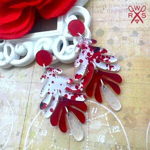 May include: Red and white leaf-shaped earrings with a splatter design. The earrings have a red circular top and a clear, leaf-shaped bottom. The earrings are hanging on a vintage map background.