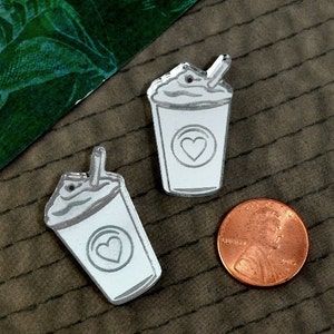 May include: Two silver-colored acrylic charms shaped like a milkshake with a straw and a heart on the side. The charms are on a brown textured surface.