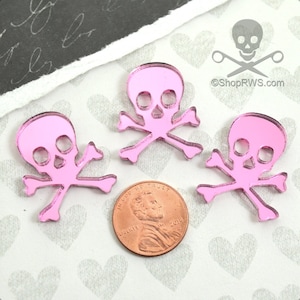 Può includere: Tre ciondoli a forma di teschio e ossa incrociate in specchio rosa. I ciondoli sono circa delle dimensioni di una moneta da 1 centesimo di euro.