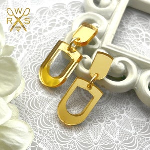 May include: A pair of gold-toned acrylic earrings with a geometric design. Each earring features a small rectangular piece connected to a larger, oval-shaped piece with a cut-out design.