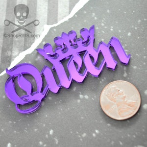 May include: A purple acrylic cutout of the word "Queen" in a stylized font, topped with a crown design. A penny is next to the cutout for size comparison. The background is a gray surface with a torn paper effect.