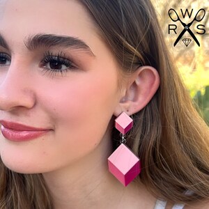Puede incluir: Un par de pendientes geométricos de color rosa y fucsia con forma cuadrada. Los pendientes son colgantes y tienen una forma cuadrada más pequeña en la parte superior.