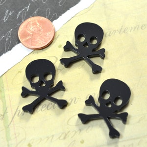 May include: Three black skull and crossbones charms are displayed on a vintage paper background. A U.S. penny is next to the charms for size comparison. The charms are made of a shiny black material.