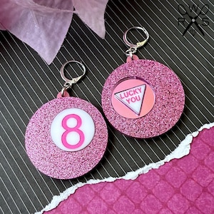 May include: Pink glitter earrings with a pink and white 8 ball charm on one earring and a pink and white triangle charm with the text "Lucky You" on the other earring.