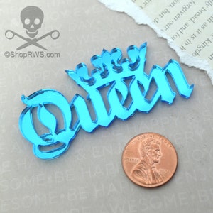 May include: A blue mirrored acrylic cutout of the word "Queen" with a crown above the word. The cutout is on a white surface with a penny for scale.