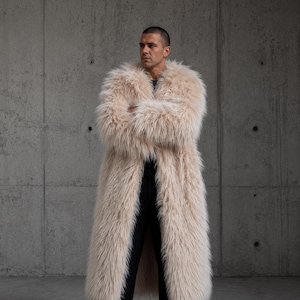 May include: A long, beige faux fur coat with a thick, shaggy texture and a wide collar. The coat is worn over a black shirt, black trousers, and black boots. The garment reaches down to the lower legs and features long sleeves with full fur coverage.