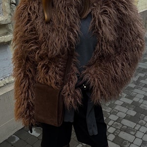 May include: A brown faux fur jacket with a shaggy texture. The jacket is paired with a dark brown crossbody bag, black shorts, and knee-high black boots. The outfit is completed with a grey top.