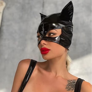 Pode incluir: Uma máscara de gato preta com orelhas pontudas. A máscara cobre os olhos e o nariz, com um acabamento brilhante. A pessoa está usando um top preto e tem batom vermelho brilhante. O fundo é cinza claro.