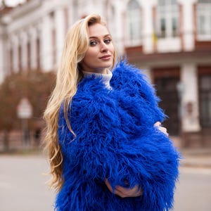 Könnte beinhalten: Ein leuchtend blauer Kunstfellmantel. Der Mantel hat eine zottelige Textur und wird über einem weißen Rollkragenpullover und einer blauen Jeans getragen. Der Hintergrund zeigt eine verschwommene Stadtlandschaft mit einem Gebäude.