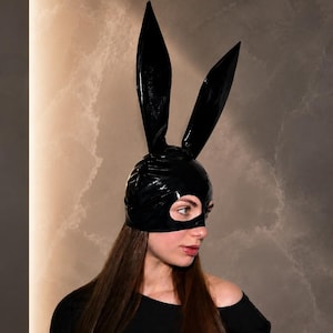 May include: A black glossy headpiece featuring two tall, pointed rabbit ears and a fitted cap design that covers the crown of the head, leaving an opening for the eyes. The material has a shiny, reflective finish and a smooth, uniform texture.