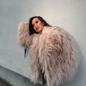 May include: A light pink faux fur coat with long, shaggy fur. The coat is worn over a grey outfit. The model is leaning against a light grey wall. The coat's texture and colour create a soft, luxurious look.