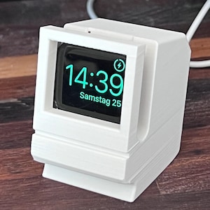 May include: White 3D printed stand shaped like an old computer with a charging dock for an Apple Watch. The watch face shows the time 14:39 and the date Samstag 25.
