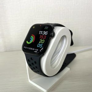 May include: A black Apple Watch Series 7 with a black sport band is on a white charging stand. The watch face displays the time 11:36, the date 153, and the day 20. The watch is connected to a white charging cable.