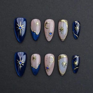 May include: Set of ten artificial nails in various designs. The nails feature a navy blue, light pink, and gold color scheme. Some nails have gold star and crescent moon accents, while others have a blue gem.