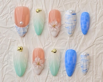 May include: A collection of ten artificial nails with various designs. Some nails are peach-colored with pearl or flower accents. Others feature blue and white patterns, or a gradient of white to mint green. Gold star and shell embellishments are also present.