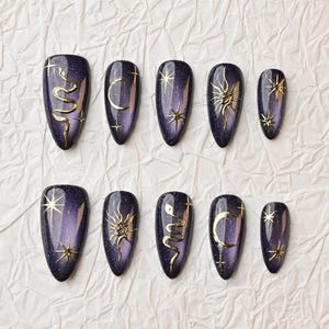 May include: A set of ten almond-shaped press-on nails featuring a deep purple, glittery base. Each nail is adorned with metallic gold decals, including designs of snakes, crescent moons, suns, and various star shapes arranged in a celestial pattern.