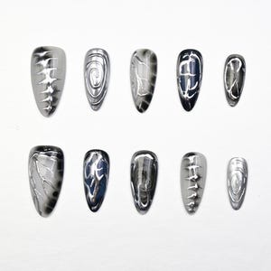 May include: A set of ten almond-shaped false nails. Each nail features a metallic silver and black design. The designs vary, including swirls, linear patterns, and marbled effects. The nails are arranged on a white surface.