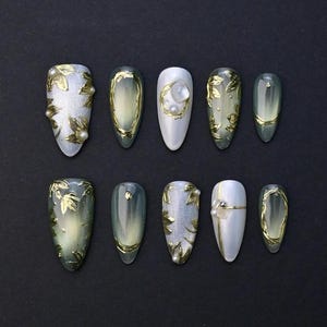 May include: A set of ten artificial nails in various shapes and designs. The nails feature gold leaf accents, pearls, and a gradient of colors, including white, silver, and green. The nails are arranged on a black surface.