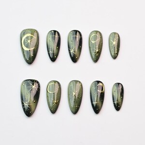 May include: Set of ten olive green press-on nails with a glossy finish. Each nail features gold celestial designs, including crescent moons and stars. The nails are arranged on a white surface.