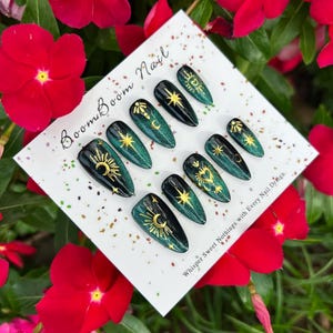 This nail set features a deep green base with intricate gold celestial designs, including stars, moons, and suns, creating a mystical and elegant look.