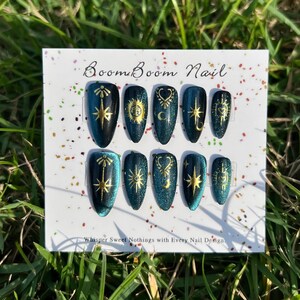 K&ouml;nnte beinhalten: Ein Set aus zehn teal- und goldfarbenen Kunstn&auml;geln. Jeder Nagel zeigt ein anderes Himmelsdesign, darunter Sterne, Monde und Sonnen. Die N&auml;gel werden auf einer wei&szlig;en Karte mit dem Text "Boom Boom Nail" und "Whisper Sweet Nothings with Every Nail Design" pr&auml;sentiert.