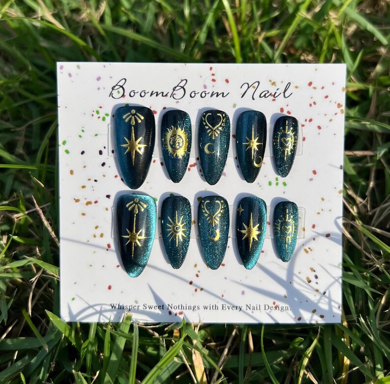 K&ouml;nnte beinhalten: Ein Set aus zehn teal- und goldfarbenen Kunstn&auml;geln. Jeder Nagel zeigt ein anderes Himmelsdesign, darunter Sterne, Monde und Sonnen. Die N&auml;gel werden auf einer wei&szlig;en Karte mit dem Text "Boom Boom Nail" und "Whisper Sweet Nothings with Every Nail Design" pr&auml;sentiert.