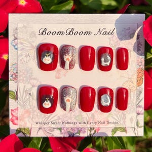 Puede incluir: Un conjunto de diez u&ntilde;as postizas rojas con un dise&ntilde;o de gato y huella de pata blanca. Las u&ntilde;as est&aacute;n dispuestas en dos filas, etiquetadas "L" y "R" para las manos izquierda y derecha. El texto "Boom Boom Nail" est&aacute; impreso en la parte superior de la tarjeta. El texto "Whisper Sweet Nothings with Every Nail Design." est&aacute; impreso en la parte inferior de la tarjeta.