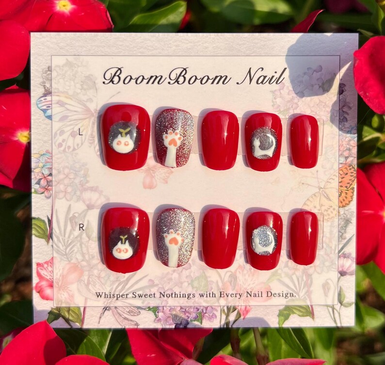 Puede incluir: Un conjunto de diez u&ntilde;as postizas rojas con un dise&ntilde;o de gato y huella de pata blanca. Las u&ntilde;as est&aacute;n dispuestas en dos filas, etiquetadas "L" y "R" para las manos izquierda y derecha. El texto "Boom Boom Nail" est&aacute; impreso en la parte superior de la tarjeta. El texto "Whisper Sweet Nothings with Every Nail Design." est&aacute; impreso en la parte inferior de la tarjeta.