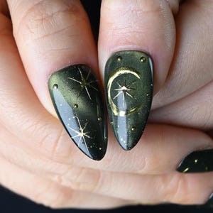 May include: Close-up of two fingernails with a dark green and gold celestial design. One nail features a gold starburst, the other a gold crescent moon and star. Small gold dots accent the design. The nails are almond-shaped.