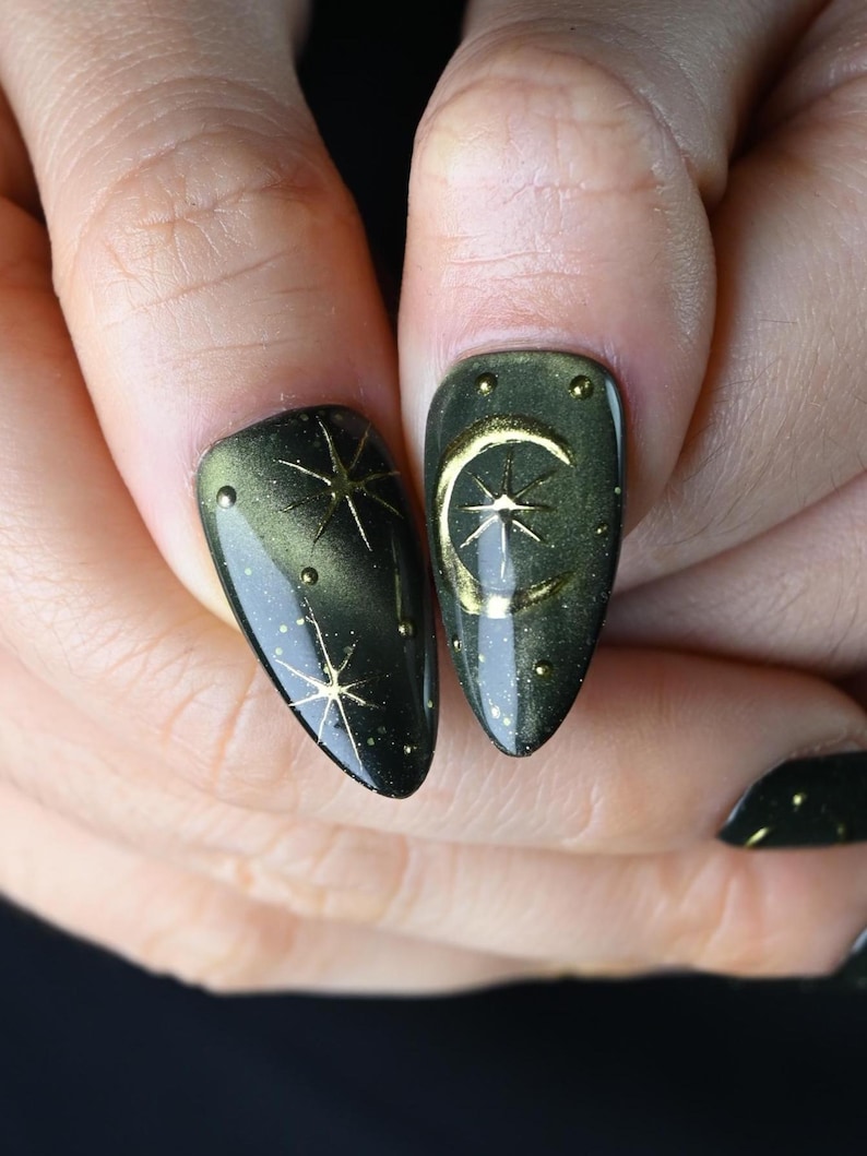 May include: Close-up of two fingernails with a dark green and gold celestial design. One nail features a gold starburst, the other a gold crescent moon and star. Small gold dots accent the design. The nails are almond-shaped.