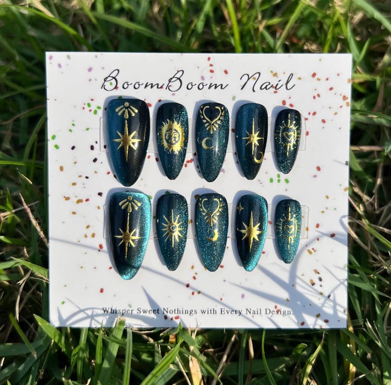 Puede incluir: Un conjunto de diez u&ntilde;as postizas verde azulado con dise&ntilde;os celestiales dorados. Las u&ntilde;as est&aacute;n dispuestas en una tarjeta blanca con el texto "Boom Boom Nail" y "Whisper Sweet Nothings with Every Nail Design".