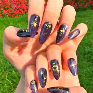 This nail design features a deep purple to black gradient base, adorned with gold celestial elements like the moon, sun, and stars, creating a luxurious and mystical vibe.