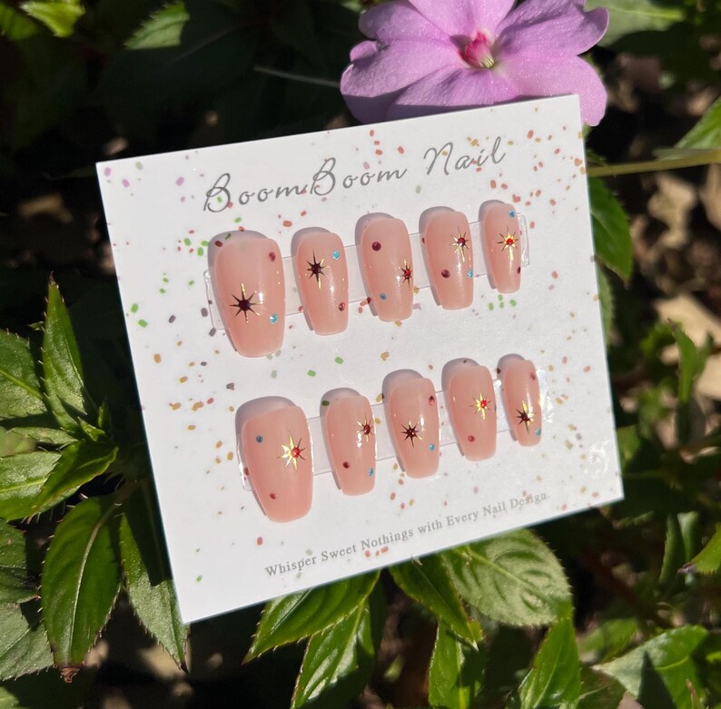 This nail set features a soft nude pink base adorned with delicate golden starbursts and colorful confetti details. Perfect for a playful yet elegant look that adds a touch of sparkle to any occasion.