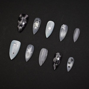 May include: A set of ten artificial nails in various shades of gray and silver. Some nails feature star designs, while others have marbled or galaxy-like patterns. The nails are almond-shaped and arranged on a black surface.