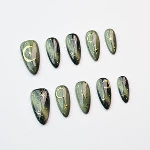 May include: A set of ten olive green press-on nails with a glossy finish. Each nail features gold celestial designs, including crescent moons and stars. The nails are oval-shaped and arranged on a white surface.