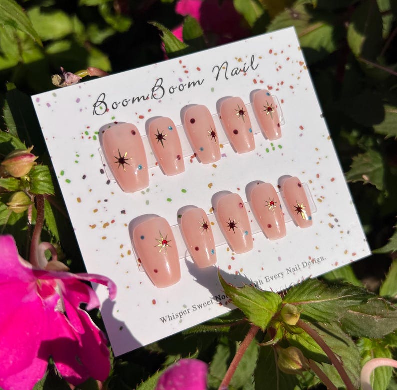 This nail set features a soft nude pink base adorned with delicate golden starbursts and colorful confetti details. Perfect for a playful yet elegant look that adds a touch of sparkle to any occasion.