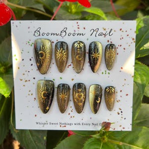 Puede incluir: Un conjunto de diez u&ntilde;as postizas con acabado de purpurina negro y dorado. Cada u&ntilde;a presenta un dise&ntilde;o dorado diferente, incluyendo lunas, estrellas y soles. Las u&ntilde;as se muestran en una tarjeta blanca con el texto "Boom Boom Nail" y "Whisper Sweet Nothings with Every Nail Done".