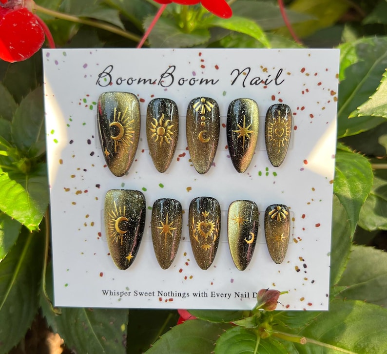 Puede incluir: Un conjunto de diez u&ntilde;as postizas con acabado de purpurina negro y dorado. Cada u&ntilde;a presenta un dise&ntilde;o dorado diferente, incluyendo lunas, estrellas y soles. Las u&ntilde;as se muestran en una tarjeta blanca con el texto "Boom Boom Nail" y "Whisper Sweet Nothings with Every Nail Done".