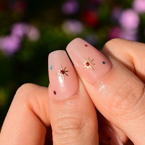 This nail set features a soft nude pink base adorned with delicate golden starbursts and colorful confetti details. Perfect for a playful yet elegant look that adds a touch of sparkle to any occasion.