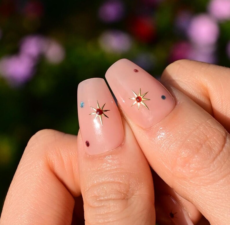 This nail set features a soft nude pink base adorned with delicate golden starbursts and colorful confetti details. Perfect for a playful yet elegant look that adds a touch of sparkle to any occasion.