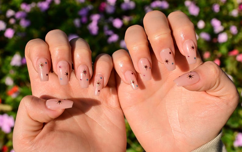 This nail set features a soft nude pink base adorned with delicate golden starbursts and colorful confetti details. Perfect for a playful yet elegant look that adds a touch of sparkle to any occasion.